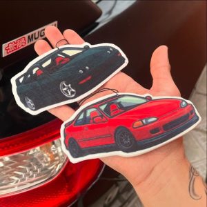 Dos ambientadores recién hechos con forma de Honda Civic e Integra, sostenidos en la mano junto a un carro